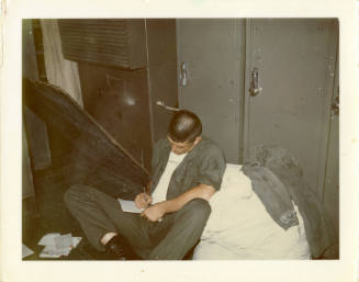 Printed color photograph of a Marine sitting in front of lockers looking down and writing a let…