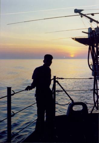 Color photograph of crew member on USS Intrepid silhouetted against a sunset over the water