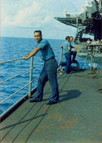 Printed color photograph of Agustin Ramos standing at the rails of USS Intrepid's hangar deck l…