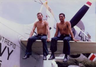 Color photograph of two sailors, shirtless, sitting the horizontal stabilizer of an airplane