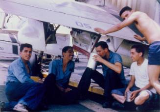 Color photograph of sailors sitting on the flight deck under a horizontal stabilizer 