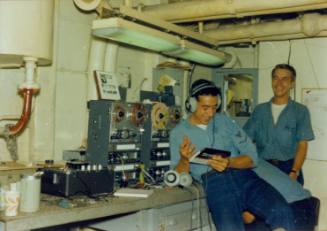 Color photograph of two sailors in a ship compartment listening to audiotapes