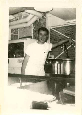 Printed black and white photograph of an enlisted sailor leaning on the spout of a vat of hot f…