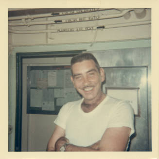 Color photograph of a man with his arms crossed smiling at the camera in the sick bay admin off…