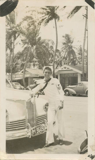 Black and white photograph of a sailor in a white uniform leaning on the front of a car, with p…