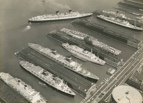 Black and white photograph of USS Intrepid docked in New York City with ocean liners at piers o…