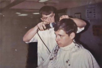 Color photograph of a barber cutting a crewmember's hair in USS Intrepid's barber shop
