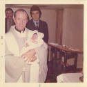 Printed color photograph of a priest holding a baby in USS Intrepid's chapel with two men stand…