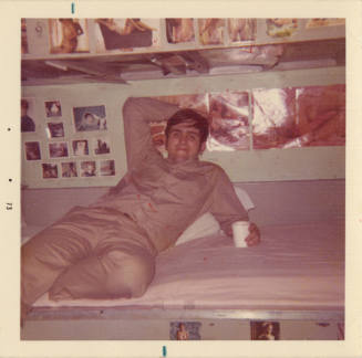 Printed color photograph of a man reclining on a bunk on board USS Intrepid