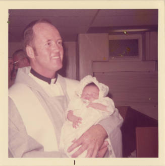 Printed color photograph of a priest holding a baby in a baptismal outfit on board USS Intrepid