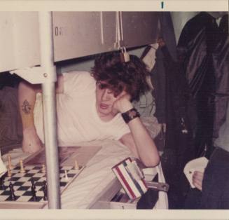 Color photograph of a man in a white t-shirt lounging on a ship's bunk and playing chess