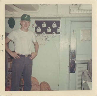 Color photograph of a man wearing a white short-sleeved shirt standing in the post office on bo…