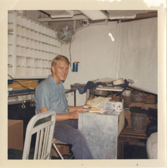 Color photograph of a man in a working dungarees uniform sitting in the post office on board US…