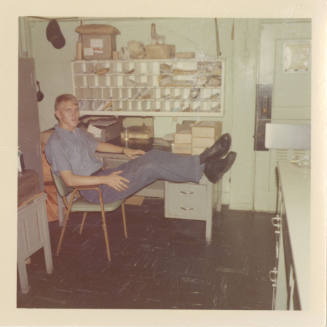 Color photograph of a sailor sitting with his feet resting on a desk in USS Intrepid's post off…