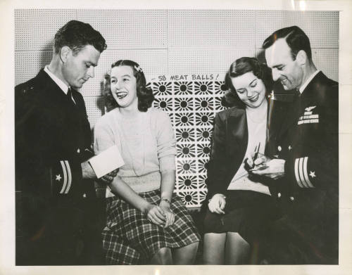 Printed black and white photograph of two officers signing autographs for two women with a scor…