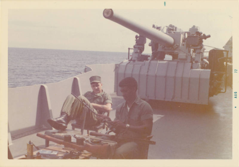 Color photograph of two Marines sitting in front of a gun mount on USS Intrepid