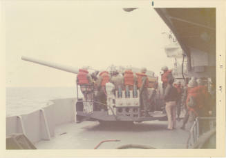 Color photograph of U.S. Marines wearing orange life vests standing on a gun mount on USS Intre…