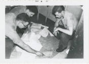 Black and white photograph of three crew members looking at plans on the floor of a ship space