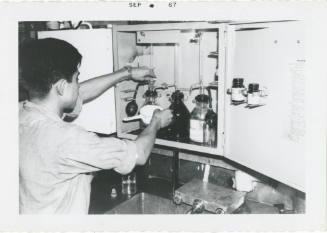 Black and white photograph of a crew member examining samples at the oil and water testing lab