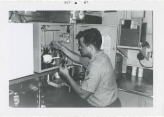 Black and white photograph of a crew member examining bottles in the oil and water testing lab