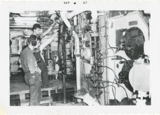 Black and white photograph of two crew members working in the lower level of a ship's fire room