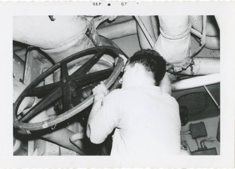 Black and white photograph of a crew member adjusting a large valve wheel in a fire room