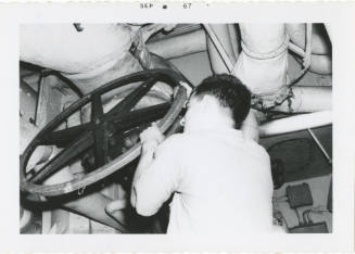 Black and white photograph of a crew member adjusting a large valve wheel in a fire room