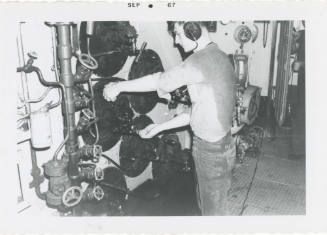 Black and white photograph of a crew member operating a fuel oil burner in a fire room