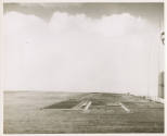 Black and whote photograph of USS Intrepid's flight deck with a large wave cresting over it wit…