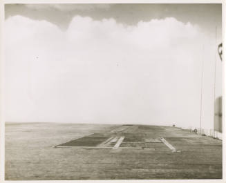 Black and whote photograph of USS Intrepid's flight deck with a large wave cresting over it wit…