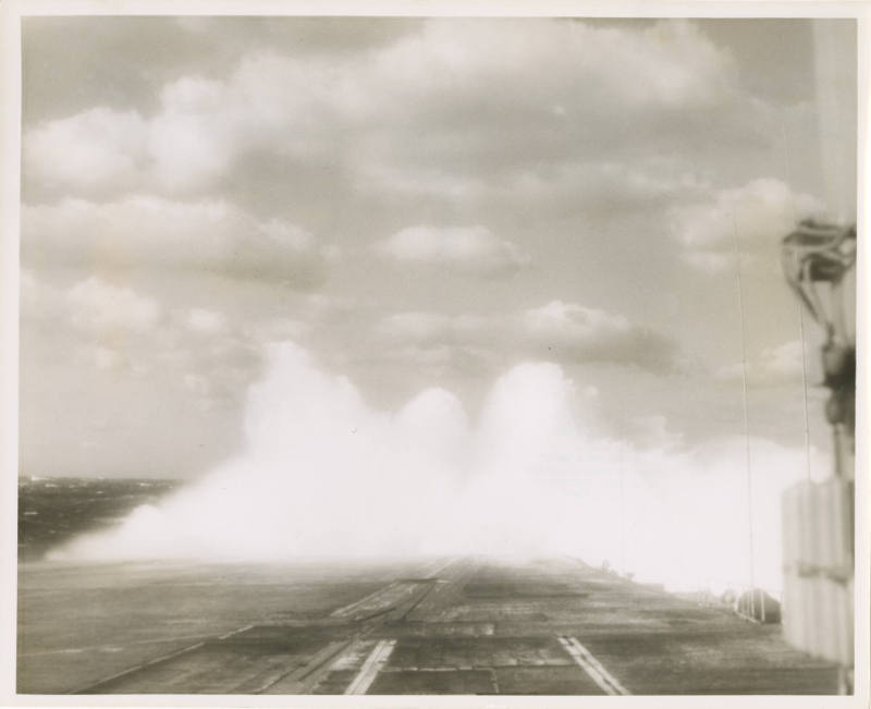 Black and white photograph of USS Intrepid's flight deck with a wave cresting over it and the s…