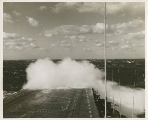 Black and white photograph of a large wave crashing over the aircraft carrier USS Intrepid's fl…