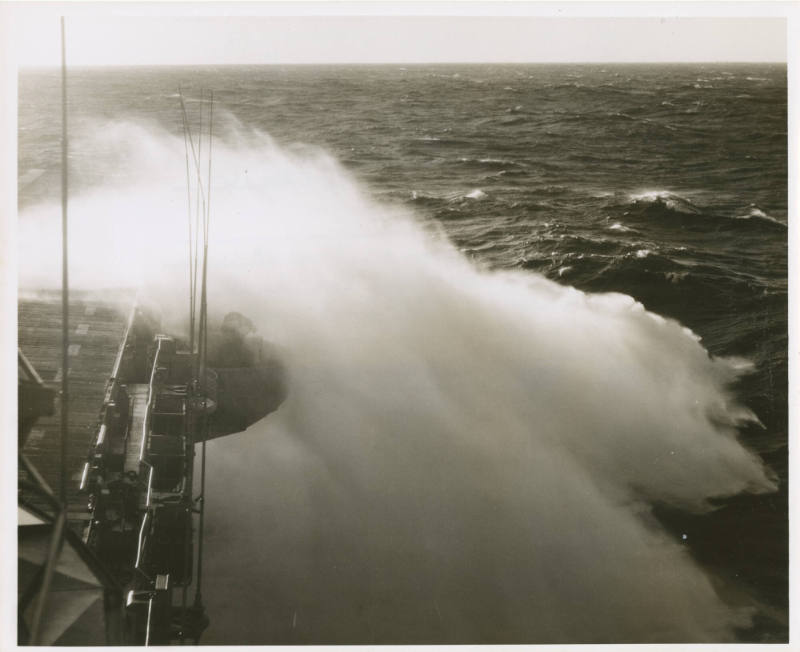 Black and white photograph of waves crashing over the aircraft carrier USS Intrepid's flight de…