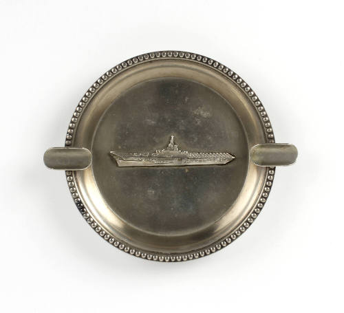 Round silver ashtray with a relief image of an aircraft carrier in center and cigarette rests o…